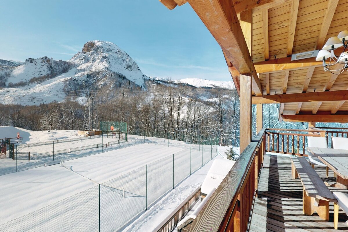 View of a snow-covered tennis court and majestic mountains from a wooden chalet balcony.