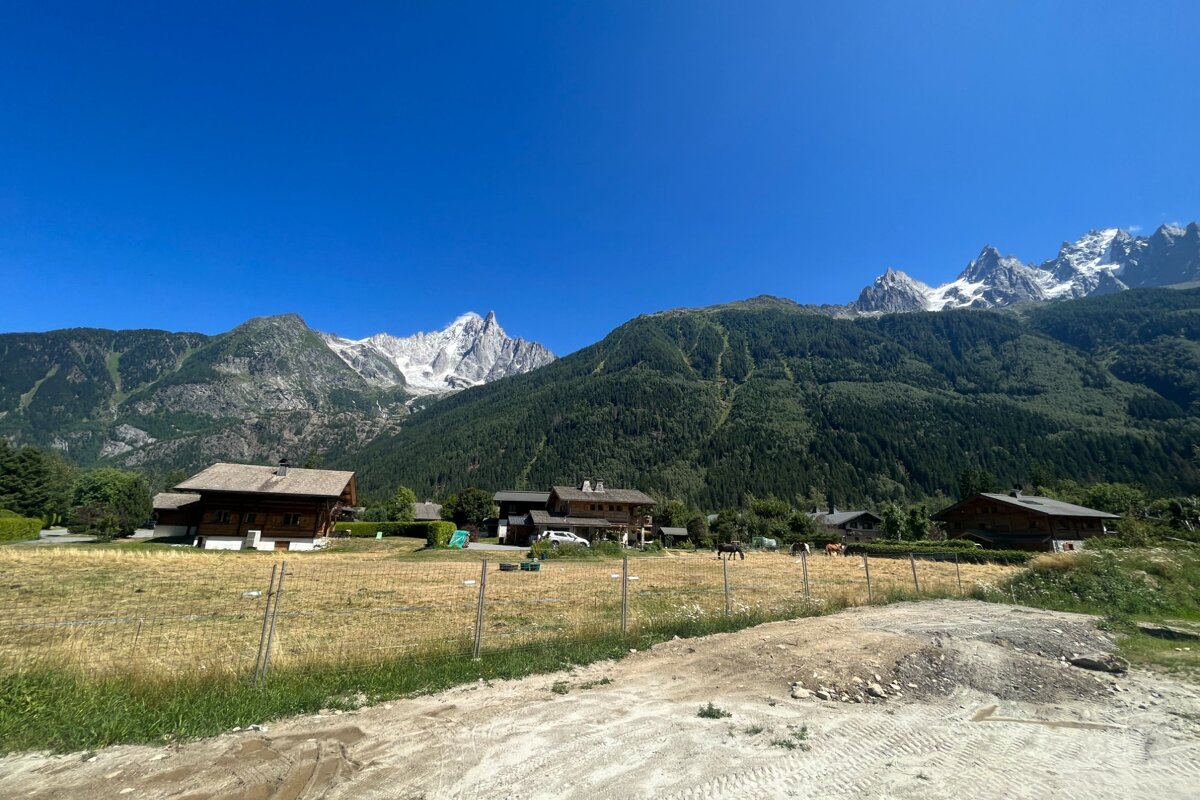 A beautiful alpine landscape features snow-capped peaks, green mountains, wooden chalets, dry fields with horses, and a clear blue sky.