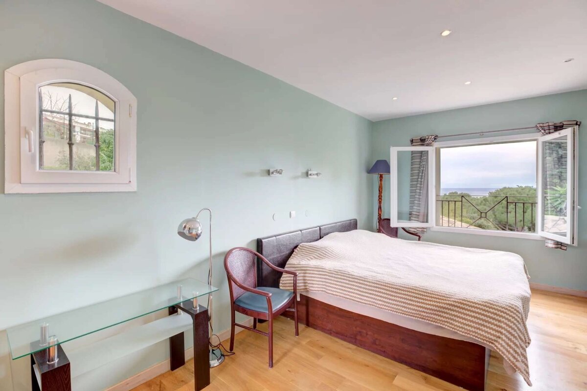 A bedroom with mint green walls, a bed, and an open window revealing a sea view and balcony. An arched window is on the left.