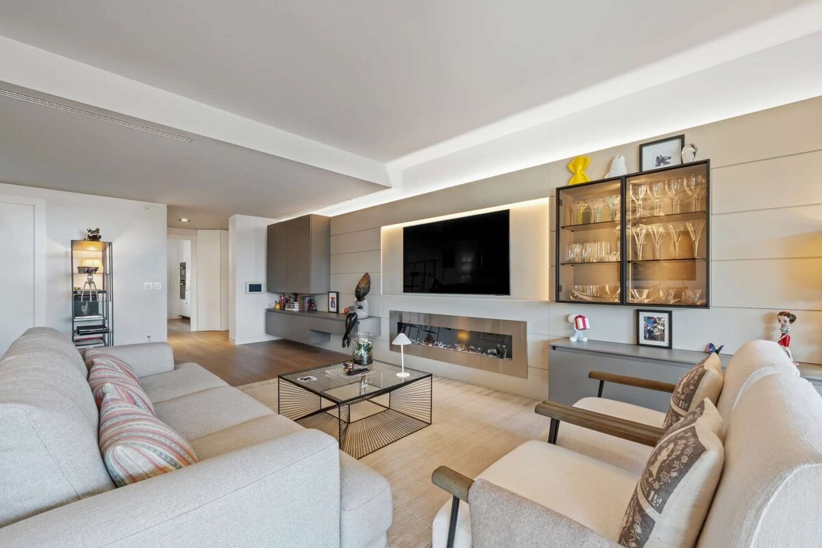A modern living room featuring a large TV over a linear fireplace, a comfortable grey sofa, armchairs, and a lighted display cabinet with glassware.