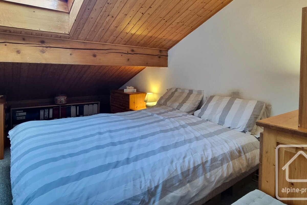 A cozy attic bedroom with a sloped wooden ceiling, exposed beams, and a bed with light gray and white striped bedding.