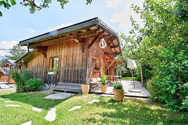 A cozy wooden cabin with an outdoor deck, potted plants, and a grassy lawn surrounded by lush green trees on a sunny day.