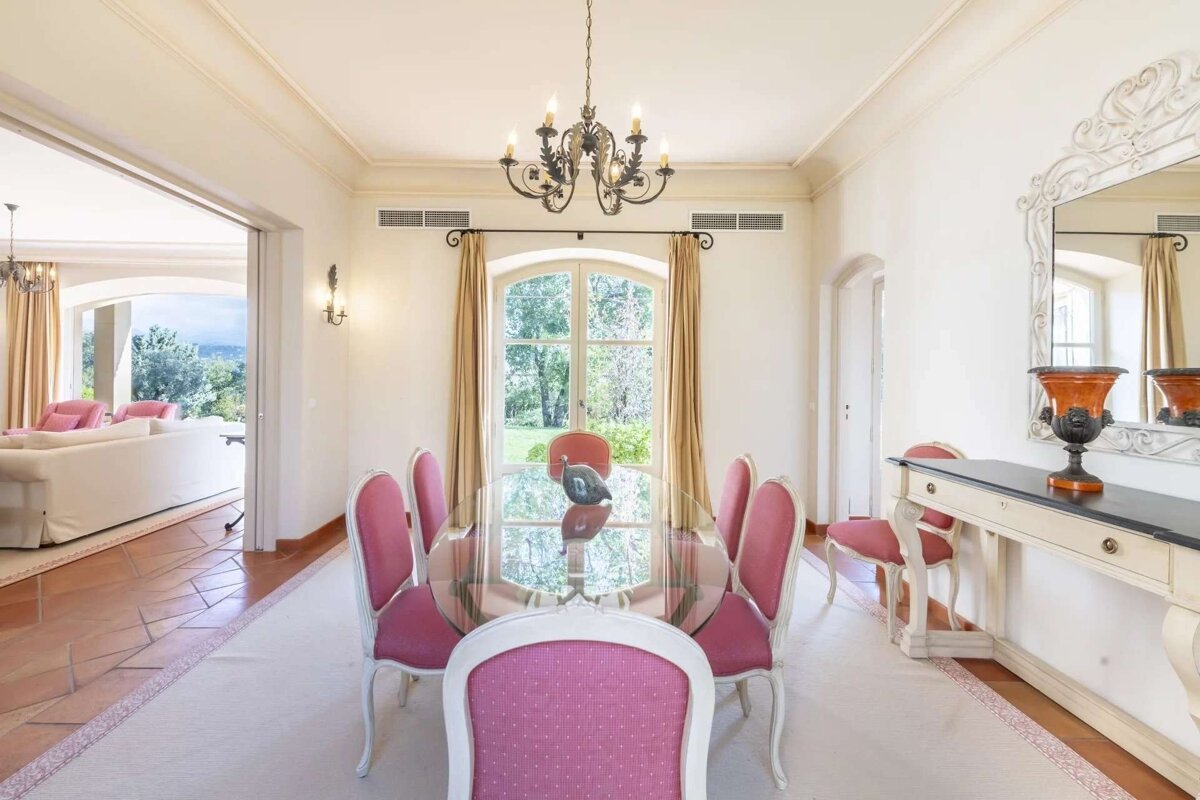 An elegant dining room featuring a glass table surrounded by pink chairs, an ornate chandelier, and a large window overlooking lush greenery. It connects to a living area.
