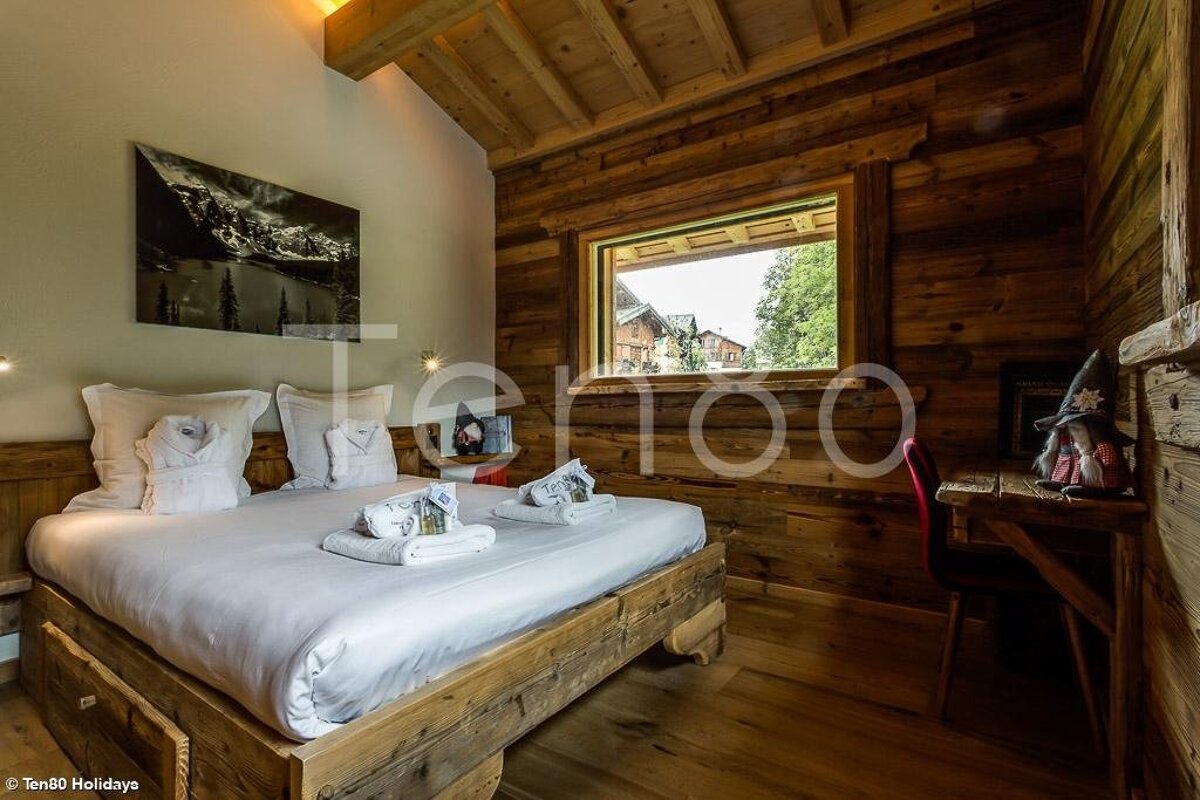 Cozy rustic cabin bedroom with a large wooden bed, white linens, wood-paneled walls, and a window overlooking outdoor chalets.