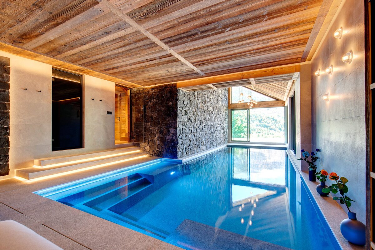 Luxurious indoor pool with wooden ceiling, stone walls, and scenic mountain views, enhanced by ambient lighting.