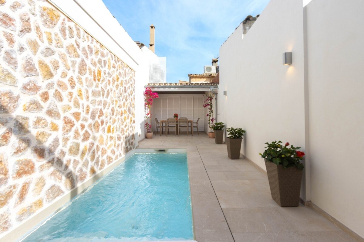 Bright, narrow courtyard with a small pool, stone wall, and dining space under a white pergola adorned with pink bougainvillea.