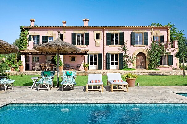 A pink house with dark shutters overlooks a sparkling blue swimming pool surrounded by a green lawn and lounge chairs on a sunny day.