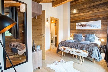 Cozy chalet bedroom with wood-paneled walls, a faux fur bedspread, a cowhide bench, and a mirror reflecting a rocking chair.