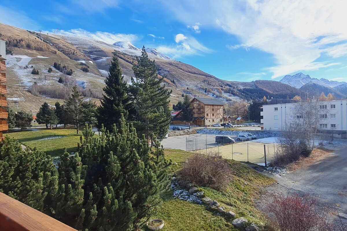 Snowy mountain village with pine trees, various buildings, and patches of snow under a bright blue sky, hinting at early winter.