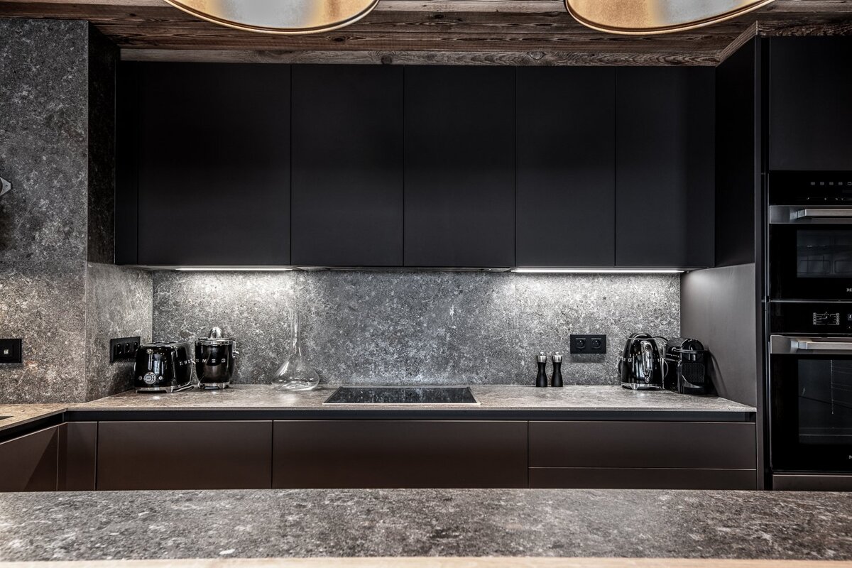 A sleek, modern kitchen featuring dark cabinets, textured gray stone counters & backsplash, illuminated by under-cabinet lighting.