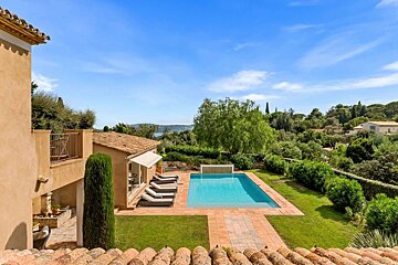 A luxurious Mediterranean villa with a sparkling blue pool, lush green gardens, and a distant sea view under a clear sky.