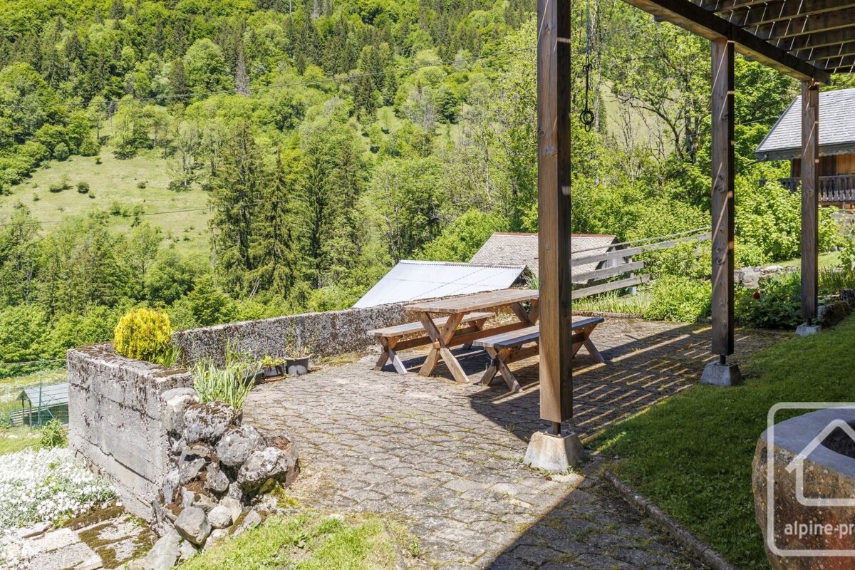 A sunny patio with a wooden picnic table overlooks a lush, green forested mountain valley with houses and outbuildings nestled amongst the trees.