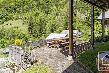 A rustic outdoor patio with a wooden picnic table under a pergola, offering views of a lush green mountain forest and distant rooftops.