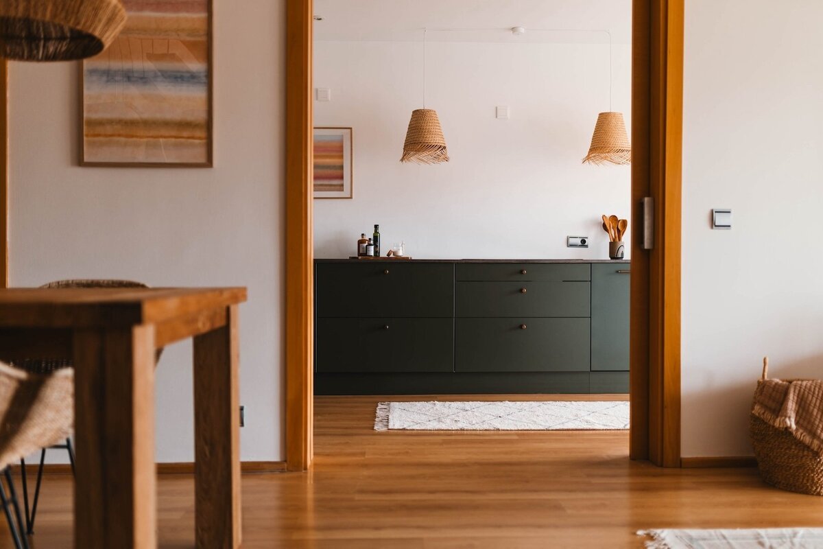 A warm interior view from a dining area, through a doorway, to a kitchen featuring dark green cabinets, wooden floors, and woven pendant lights.