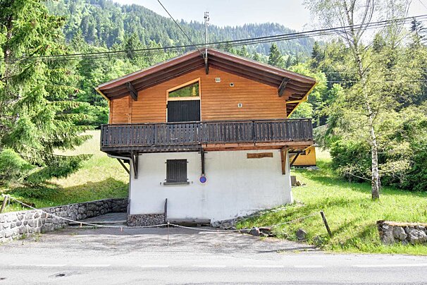 A two-story chalet, with a wooden upper floor and balcony over a white lower floor, sits amidst a lush green mountain landscape by a paved road.