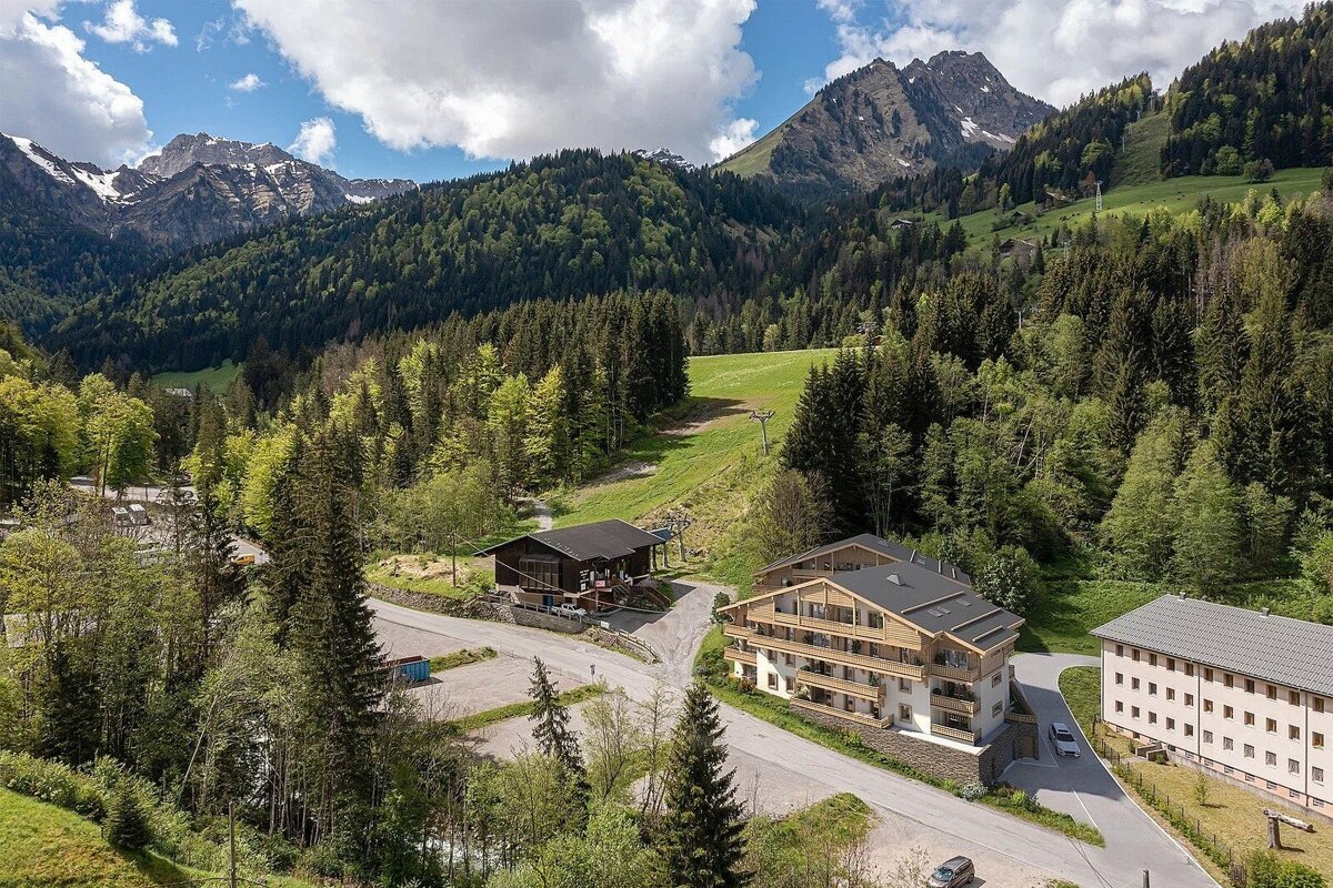 Aerial view of a mountain village with modern chalets and traditional buildings nestled in a lush green valley, surrounded by vast forests and snow-capped peaks.