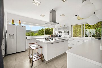 A bright, modern white kitchen with a central island, stainless steel hood, rolling cart, and large windows showcasing scenic outdoor views.