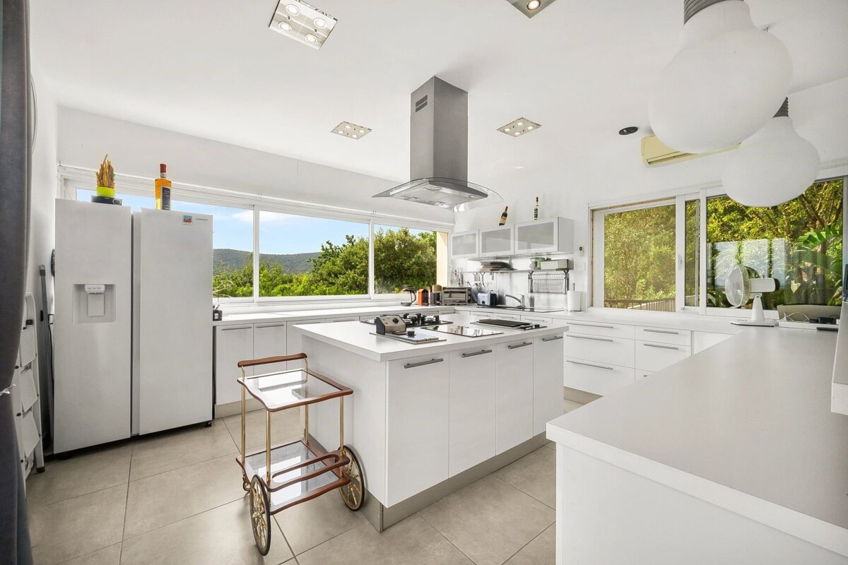 A bright, modern white kitchen featuring a central island, stainless steel hood, two-door refrigerator, and large windows with a view.