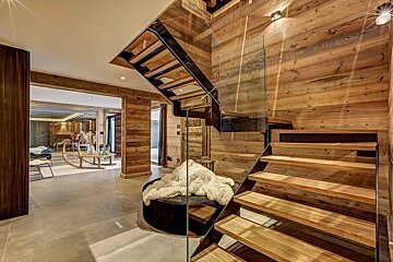 Modern chalet interior with warm wooden walls, sleek glass and wood staircase, and a cozy seating area with a fur throw.