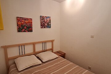 A simple bedroom with a wooden bed, striped duvet, two pillows, and two vibrant floral art prints on light walls. A small nightstand is also visible.
