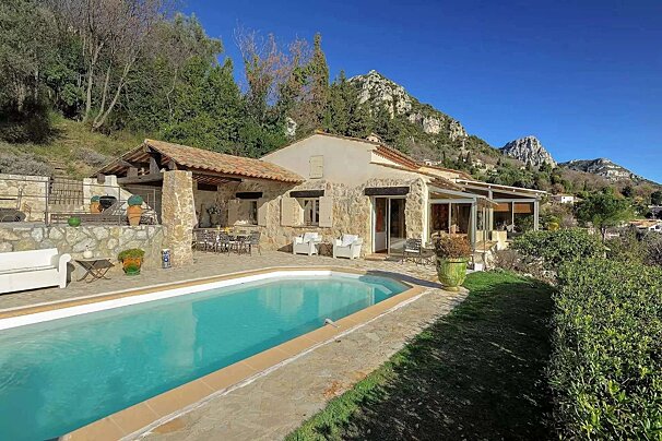 A Mediterranean stone house with a pool, nestled on a hillside with impressive mountains in the background under a clear blue sky.
