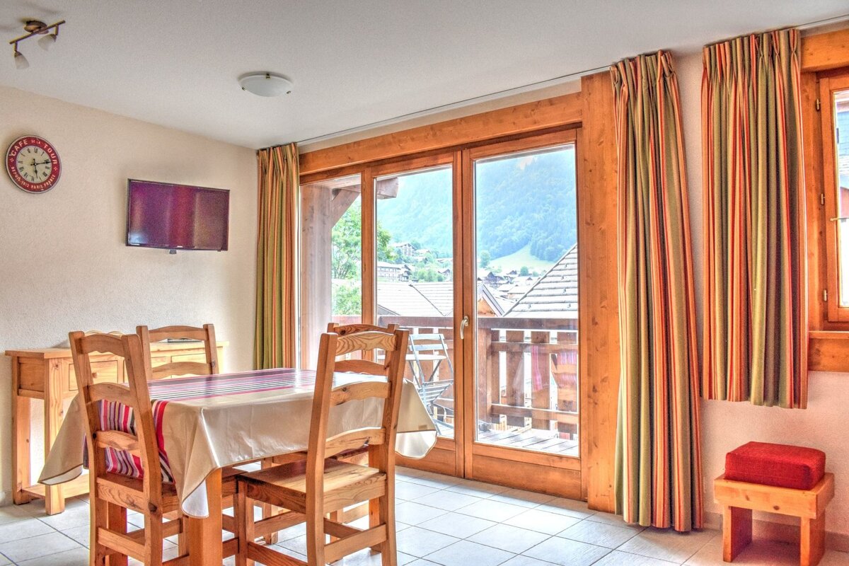 A dining room with a wooden table, chairs, wall-mounted TV, and clock. Glass doors open to a balcony with a view of a mountain village and peaks.