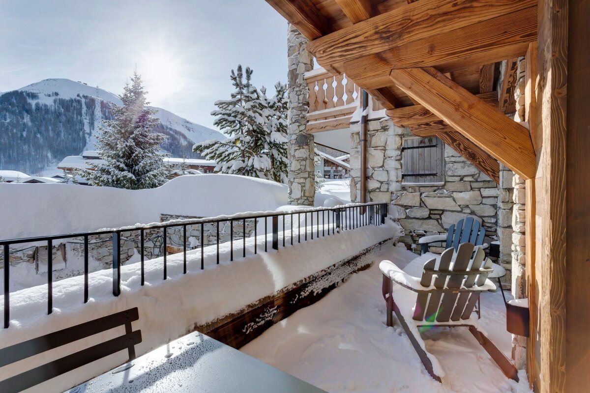 A sunny winter scene from a snowy chalet balcony. Mountains, pine trees, and snow-laden Adirondack chairs fill the picturesque view.