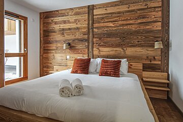 A cozy bedroom featuring a rustic wooden plank wall behind a white bed with orange pillows and rolled towels, illuminated by a large window.
