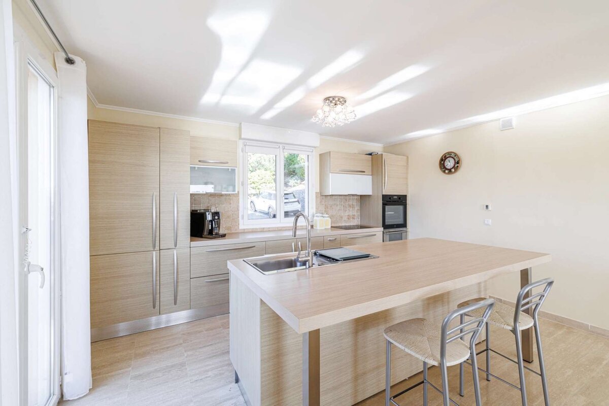 A bright, modern kitchen with light wood cabinets, a central island with a sink, and two bar stools. Sunlight streams through windows.