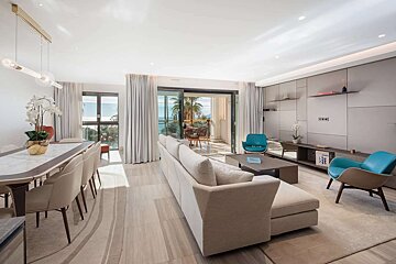 A bright, modern living/dining room with large windows, a beige sectional sofa, teal armchairs, and a balcony offering sea views.