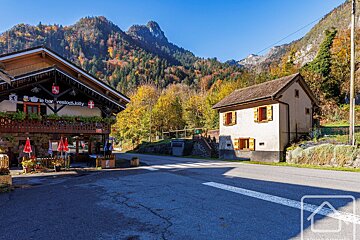 Sunny alpine village with a bar/restaurant and house, backed by mountains vibrant with autumn foliage, and a road with a crosswalk.