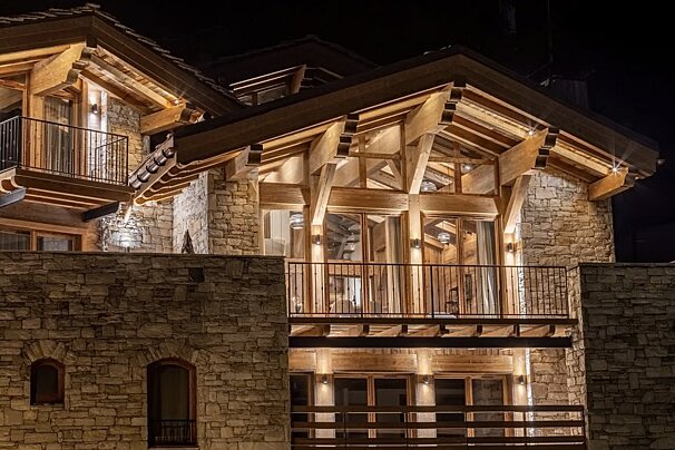 A grand, illuminated chalet at night, showcasing rich stone masonry and prominent wooden beams. Balconies and large windows add to its inviting warmth.