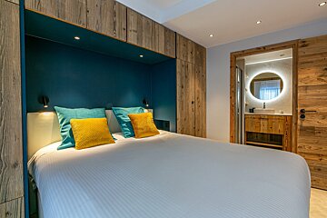 Modern bedroom with a white bed, teal and yellow pillows, wooden built-ins, and a view into a bathroom with a round backlit mirror.