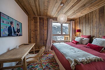 A rustic, wood-paneled bedroom features a red bed, colorful rug, and a TV. A large window offers a snowy mountain view, creating a cozy winter retreat.