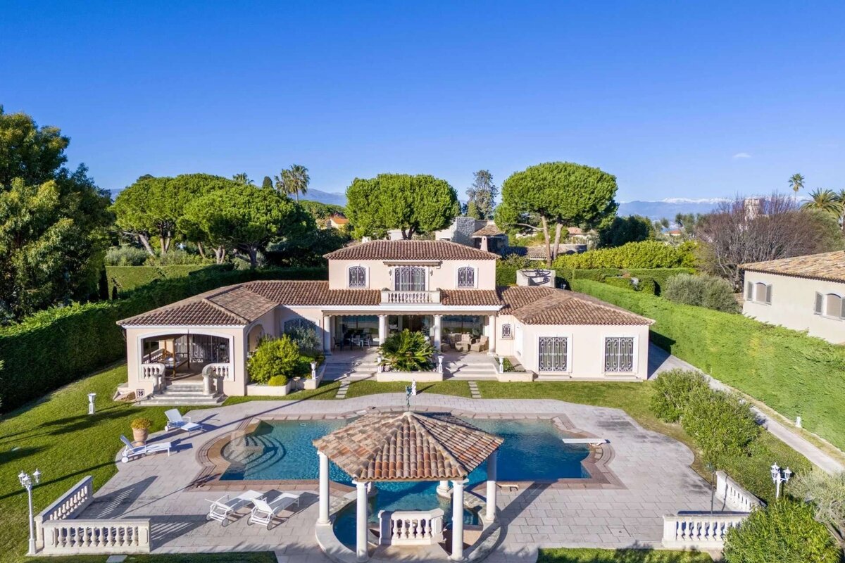 Luxurious villa with a large swimming pool, elegant gazebo, and manicured gardens, set against a backdrop of distant mountains under a blue sky.