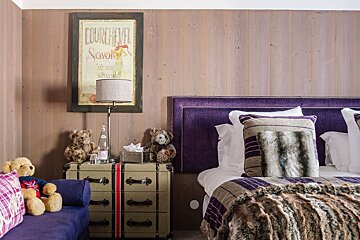 Cozy bedroom with wood paneling, a purple bed, fur blanket, luggage nightstand, teddy bears, and a vintage Courchevel poster.