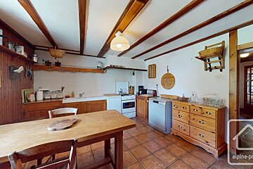 A charming, rustic kitchen with wooden ceiling beams, white walls, and a tiled floor. It features a large wooden table, stove, sink, and dishwasher.