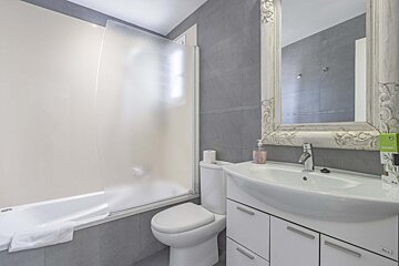 A modern bathroom with grey tiled walls, featuring a bathtub with a frosted screen, toilet, white vanity sink, and an ornate mirror.