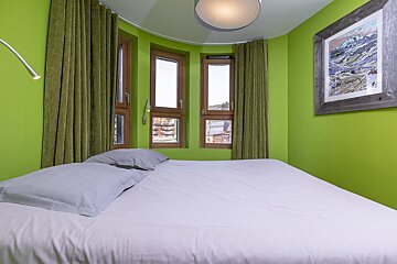 A modern bedroom with vibrant lime green walls, a large bed with grey bedding, and a curved wall featuring curtained windows. Mountain art hangs on the wall.