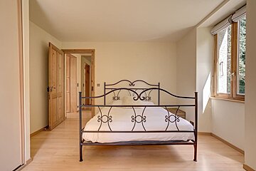 Minimalist bedroom featuring a wrought-iron bed with white linens, light wooden floors, an open door, and a window.