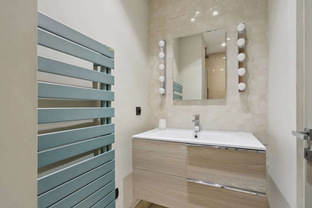 Modern bathroom features a light blue towel warmer, a wooden vanity with a sink, and a lighted mirror against a tiled wall.