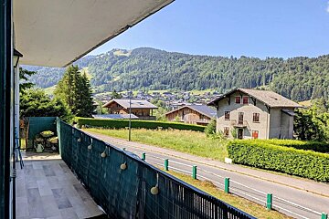 Balcony view of a mountain village with wooden chalets, lush green hills, a road, and forested mountains under a clear blue sky.