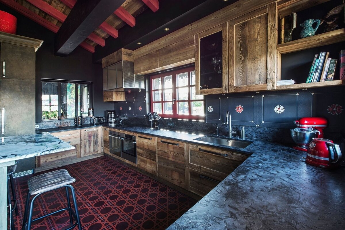 A chic kitchen with dark walls, rustic wood cabinets, dark counters, a red window frame, and a bold red patterned rug. Red ceiling beams add character.