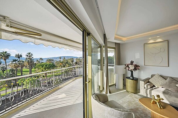 An elegant living room opens to a sunny balcony with an awning, overlooking a palm-lined coast, a road, and a sparkling bay.