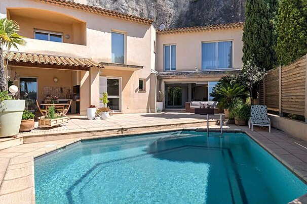 Sunny Mediterranean-style villa with a sparkling pool and multiple patios, nestled against a rocky cliff, ideal for outdoor living.