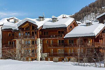Multi-story wooden buildings blanketed in heavy snow, with balconies and chimneys, under a bright blue sky in a snowy mountain landscape.