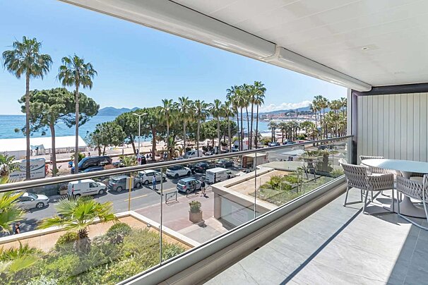 A modern balcony overlooks a vibrant coastal scene with a road, palm trees, a sandy beach, and the blue sea under a sunny sky.