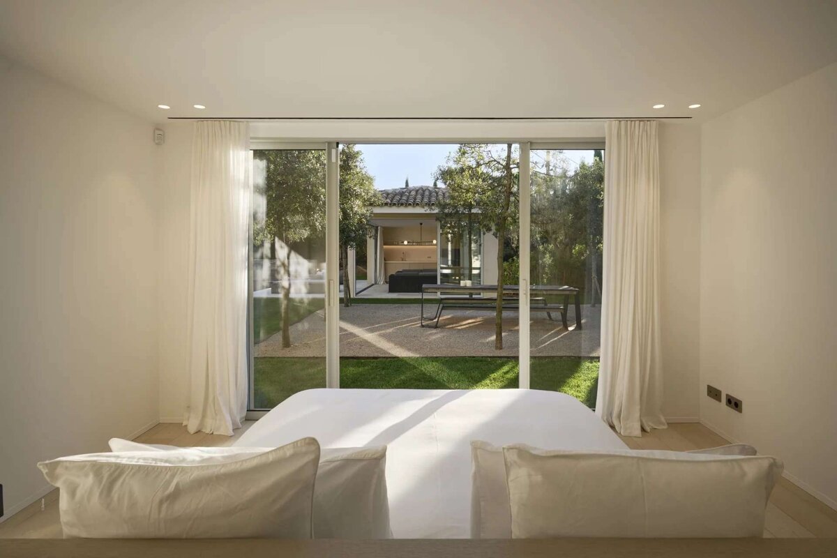 A clean, modern bedroom features a white bed facing large glass doors, opening to a lush garden with trees, gravel, and an outdoor seating area.