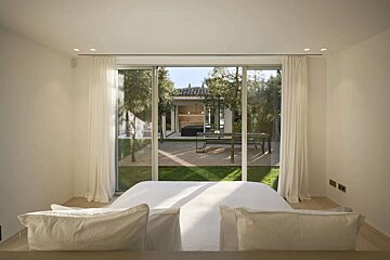 A clean, modern bedroom features a white bed facing large glass doors, opening to a lush garden with trees, gravel, and an outdoor seating area.