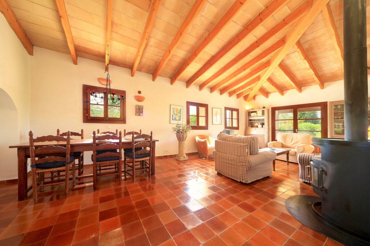 A spacious rustic room features a wooden beamed ceiling, terracotta floor, a dining area, a cozy lounge with sofas, and a large wood-burning stove.